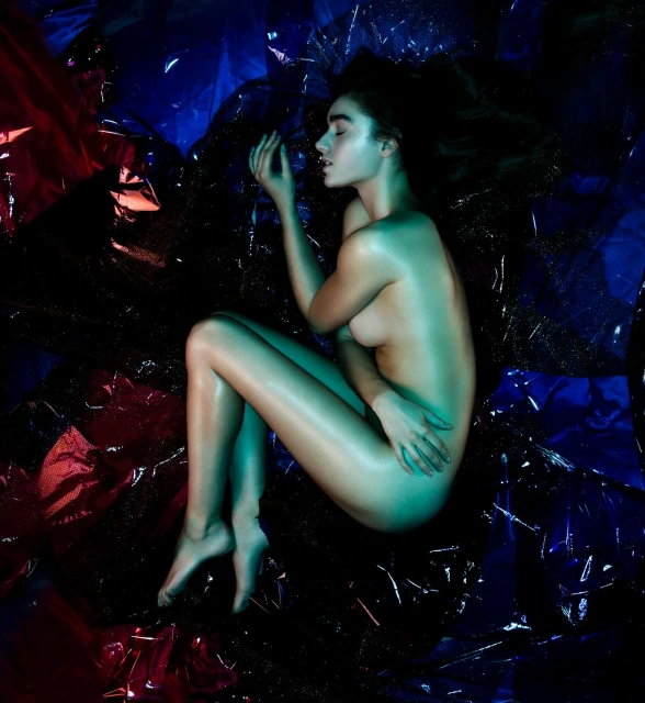 Nude woman with wet hair poses on shiny black and red cellophane with blue and green lighting in a studio photography setting.