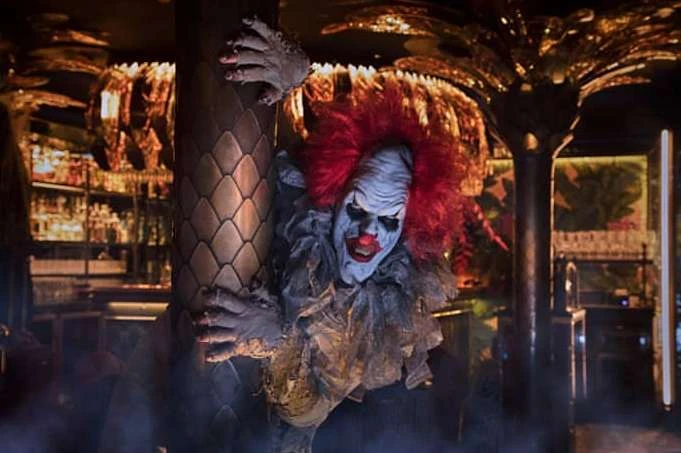 Scary clown with red hair and painted face clinging to a pillar inside a bar with tropical decor, creating a spooky atmosphere.