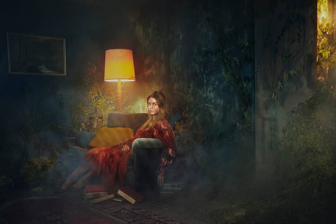 A woman in a red dress seated on a velvet armchair in an overgrown moody room lit by a glowing floor lamp
