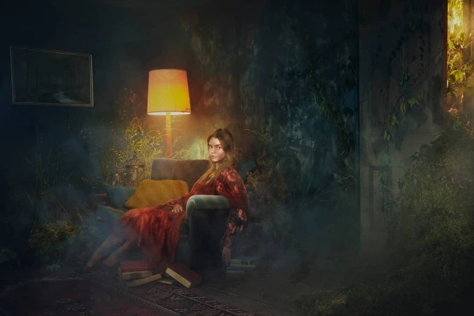 A woman in a red dress seated on a velvet armchair in an overgrown moody room lit by a glowing floor lamp