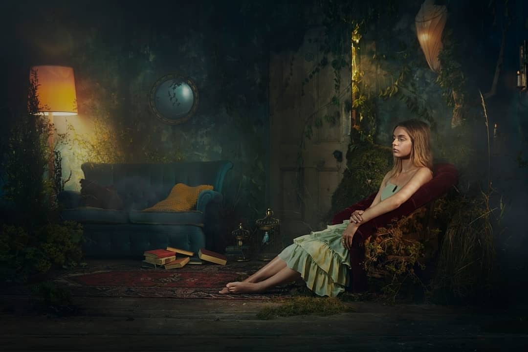 A young woman in a light green dress sits in a chair in a dimly lit room with a lamp, sofa, books, and overgrown plants.