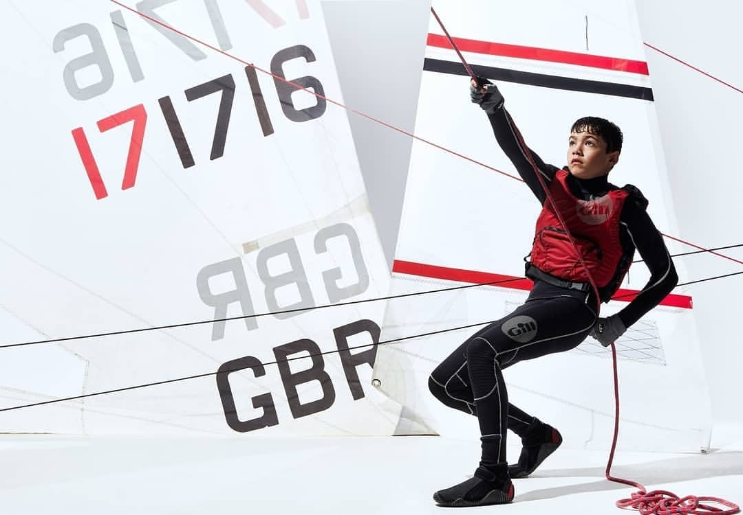 Young sailor in wetsuit and red life vest pulls a line on a sailboat with the flag of Great Britain against a white background.