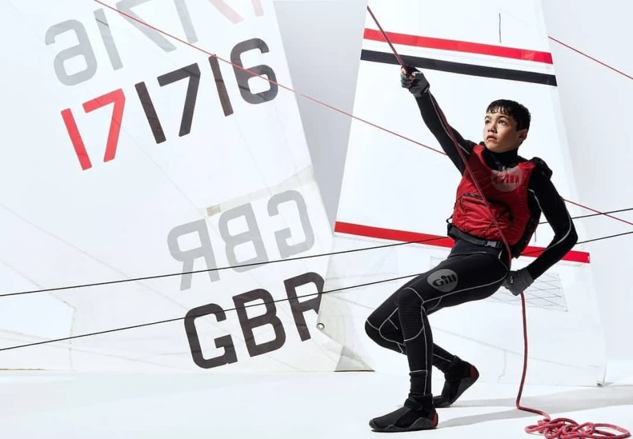 Young sailor in wetsuit and red life vest pulls a line on a sailboat with the flag of Great Britain against a white background.