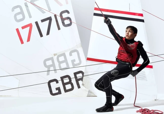 Young sailor in wetsuit and red life vest pulls a line on a sailboat with the flag of Great Britain against a white background.