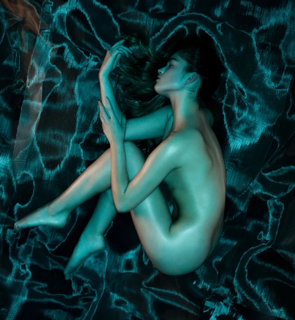 A nude fair-skinned woman poses on a draped fabric with light blue lighting, creating an artistic and sensual studio portrait.