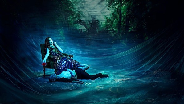 Artistic studio shot of a woman in a dress reclining in a chair with a man lying at her feet, set against a jungle background.