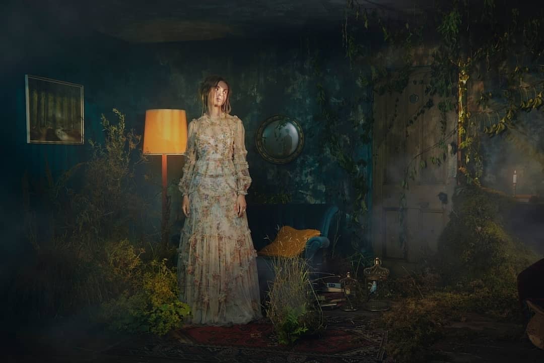 A woman in a floral dress stands in a dimly lit, overgrown interior with a lamp, plants, and antique furniture creating a vintage aesthetic.