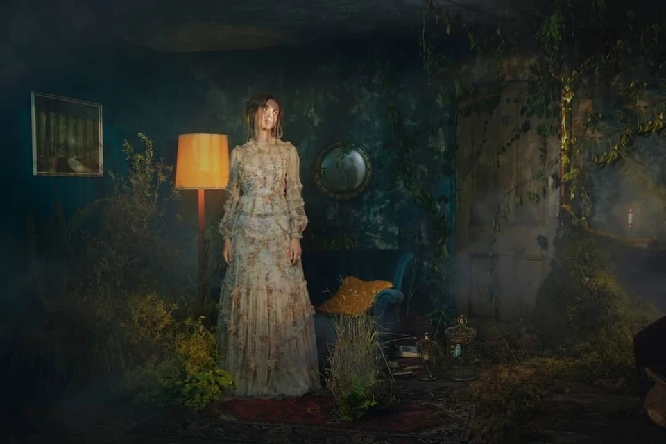A woman in a floral dress stands in a dimly lit, overgrown interior with a lamp, plants, and antique furniture creating a vintage aesthetic.