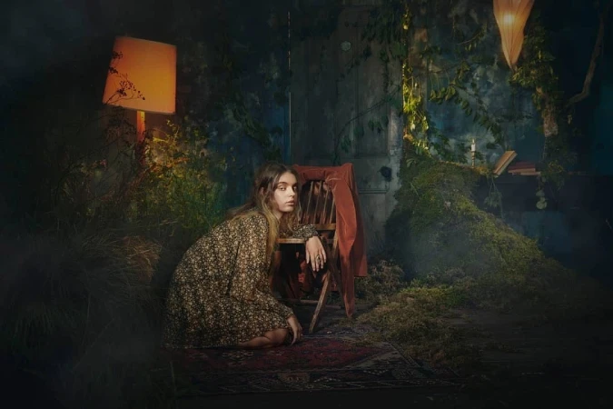 Young woman in floral dress sits near a rustic chair indoors surrounded by foliage and a warm lamp providing soft light.