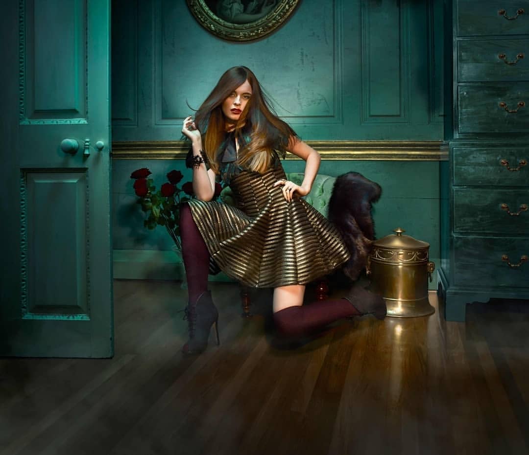 A fashionable woman in a gold-striped dress poses in an elegant interior with dark green walls, wooden floors, a wardrobe, and roses.