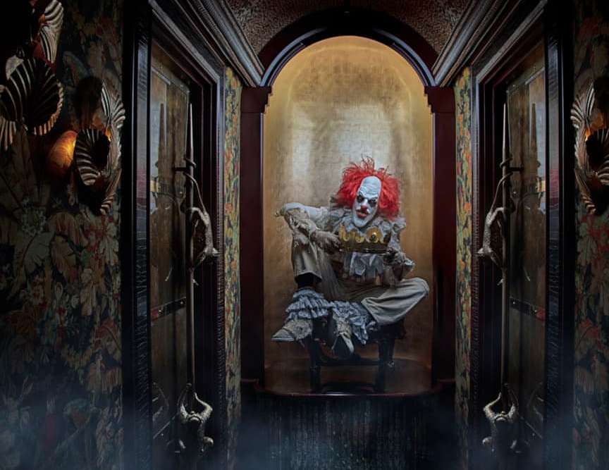 Creepy clown with orange hair sits holding golden crown in ornate corridor with gold accents and floral wallpaper in moody lighting.