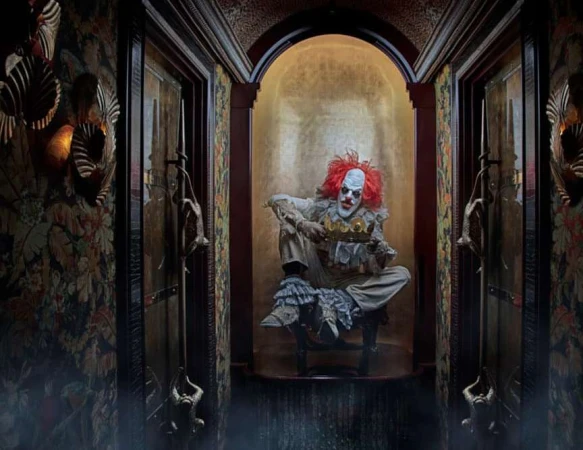 Creepy clown with orange hair sits holding golden crown in ornate corridor with gold accents and floral wallpaper in moody lighting.