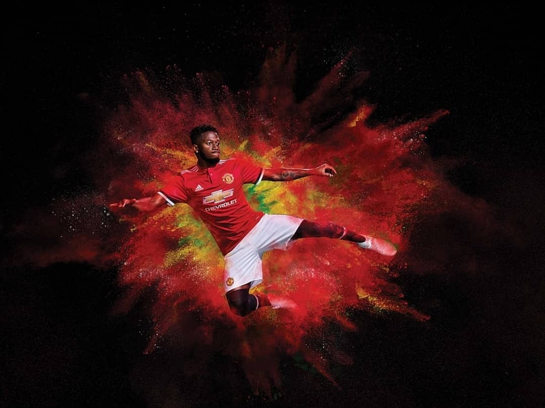 A soccer player in mid-air, poised to kick, wearing a Manchester United jersey, set against a red, yellow, and black backdrop.