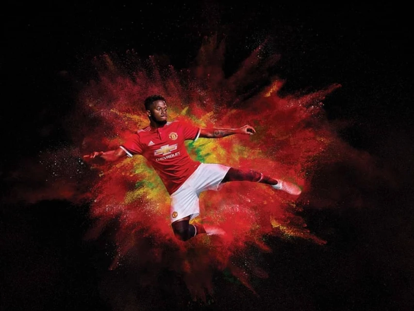 A soccer player in mid-air, poised to kick, wearing a Manchester United jersey, set against a red, yellow, and black backdrop.