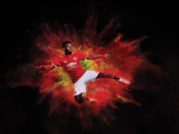 A soccer player in mid-air, poised to kick, wearing a Manchester United jersey, set against a red, yellow, and black backdrop.
