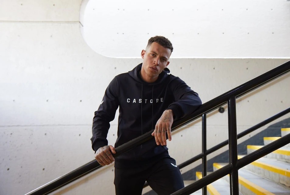 Athletic adult male in a hooded sweatshirt poses on a staircase with concrete walls, illuminated by natural light indoors.