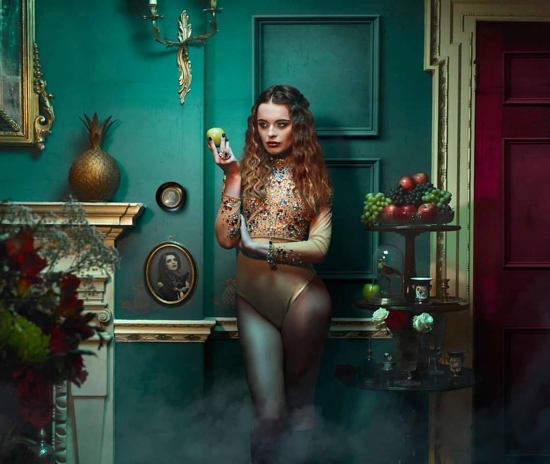 Woman in gold bodysuit holds green apple in indoor portrait with dark teal walls, gold accents, fruit and decorative objects.