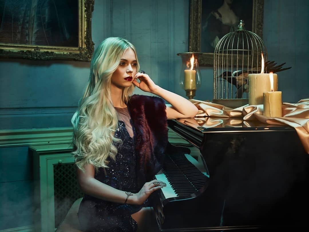 Young woman with long blonde hair and a dark dress sits at a piano next to candles and a birdcage in a dimly lit, ornate room.