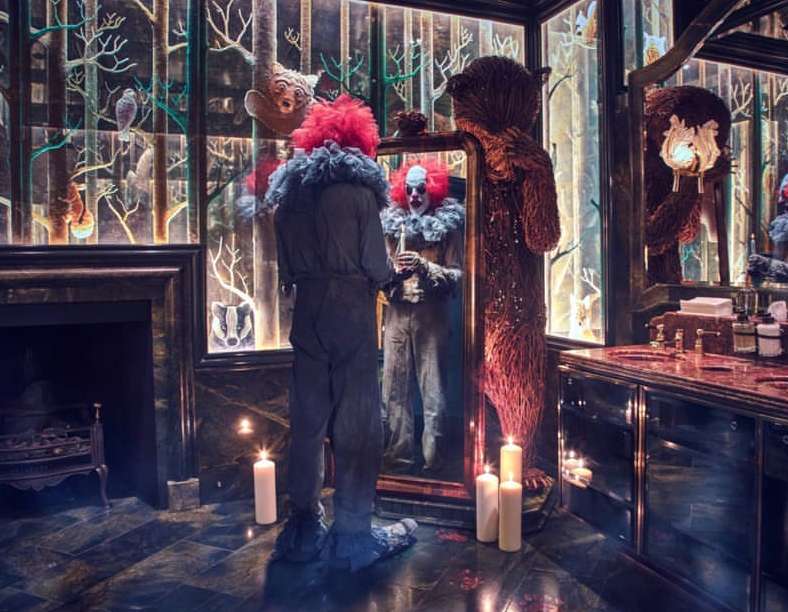 Person in clown costume stands before a mirror in a dimly-lit, classically-designed bathroom with a large decorative bear.