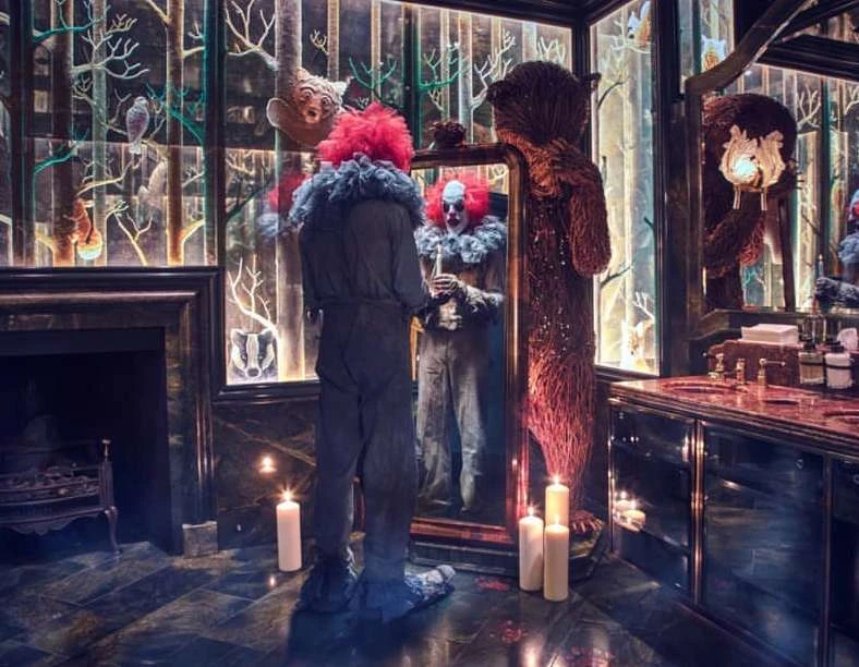 Person in clown costume stands before a mirror in a dimly-lit, classically-designed bathroom with a large decorative bear.