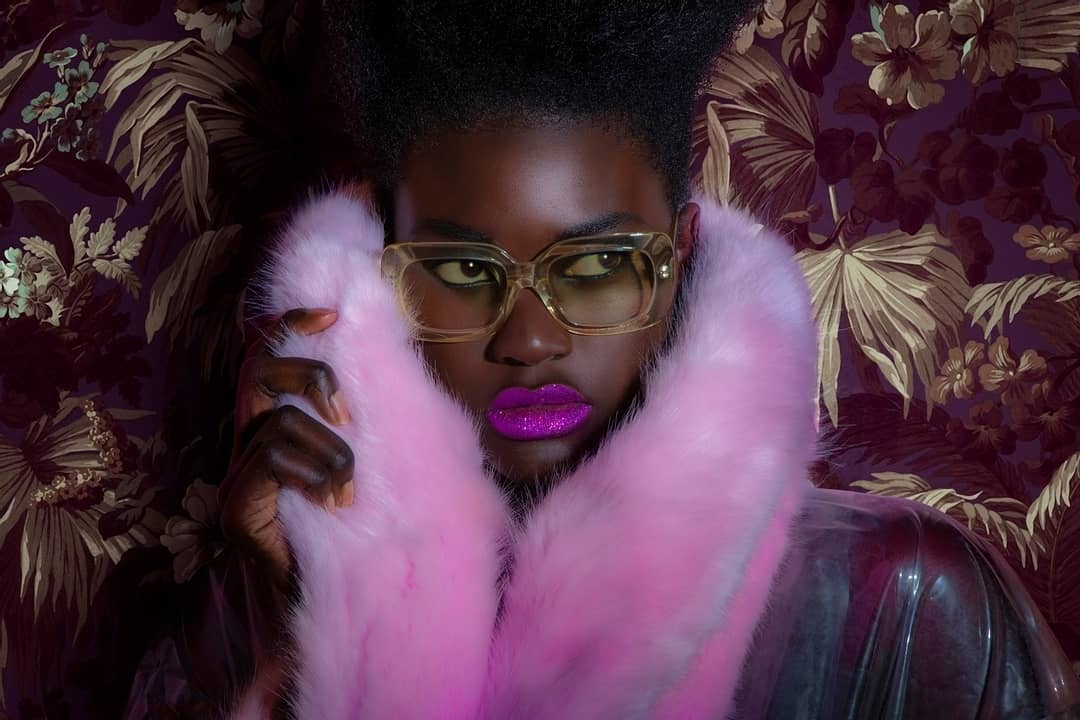 Close-up of an African-American woman with bright pink lipstick, wearing a pink fur stole and clear glasses, against a floral background.