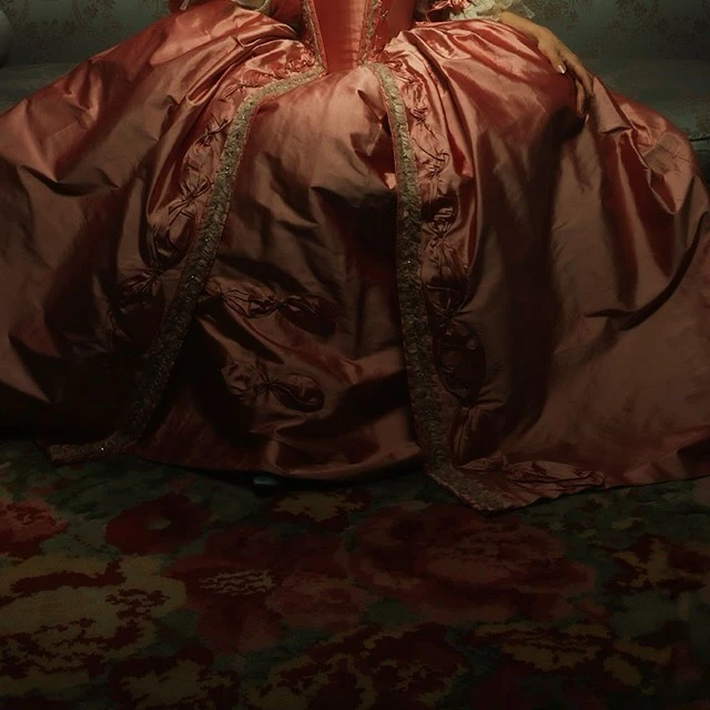 Close-up of a ruffled, coral-colored ballgown with lace trim and ribbon details, covering a patterned floral rug indoors.