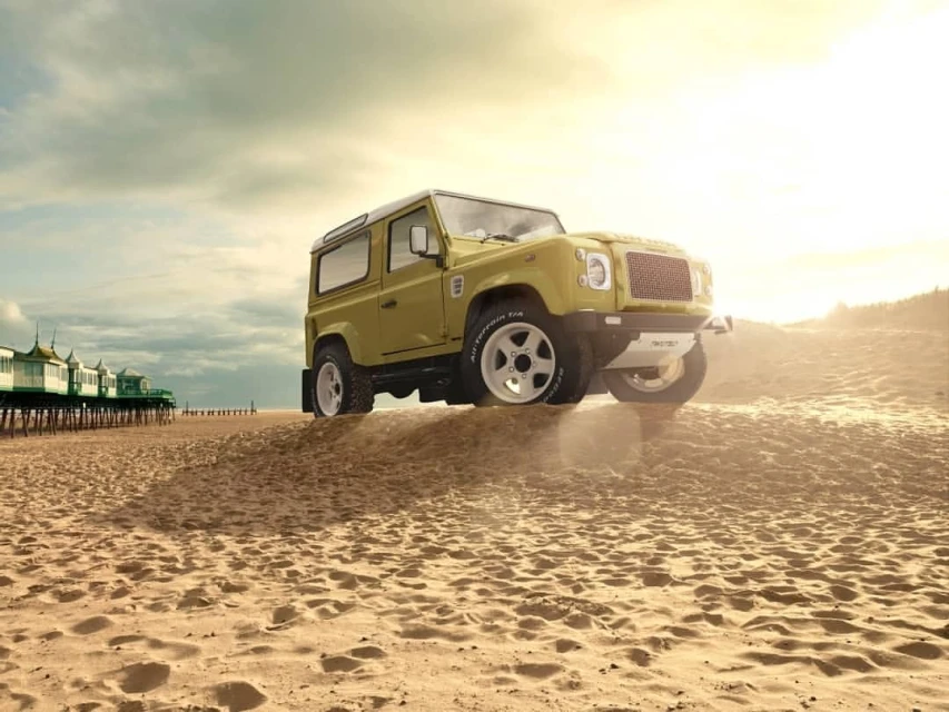 Off-road vehicle parked on a sandy beach with a distant pier under a cloudy sky at sunset. Vehicle is gold and white.