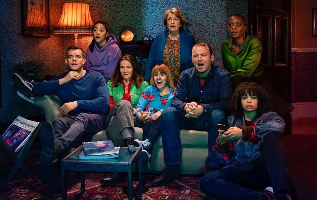 A group of eight diverse adults are gathered indoors, exhibiting a variety of expressions while seated in a warmly lit room with patterned wallpaper.