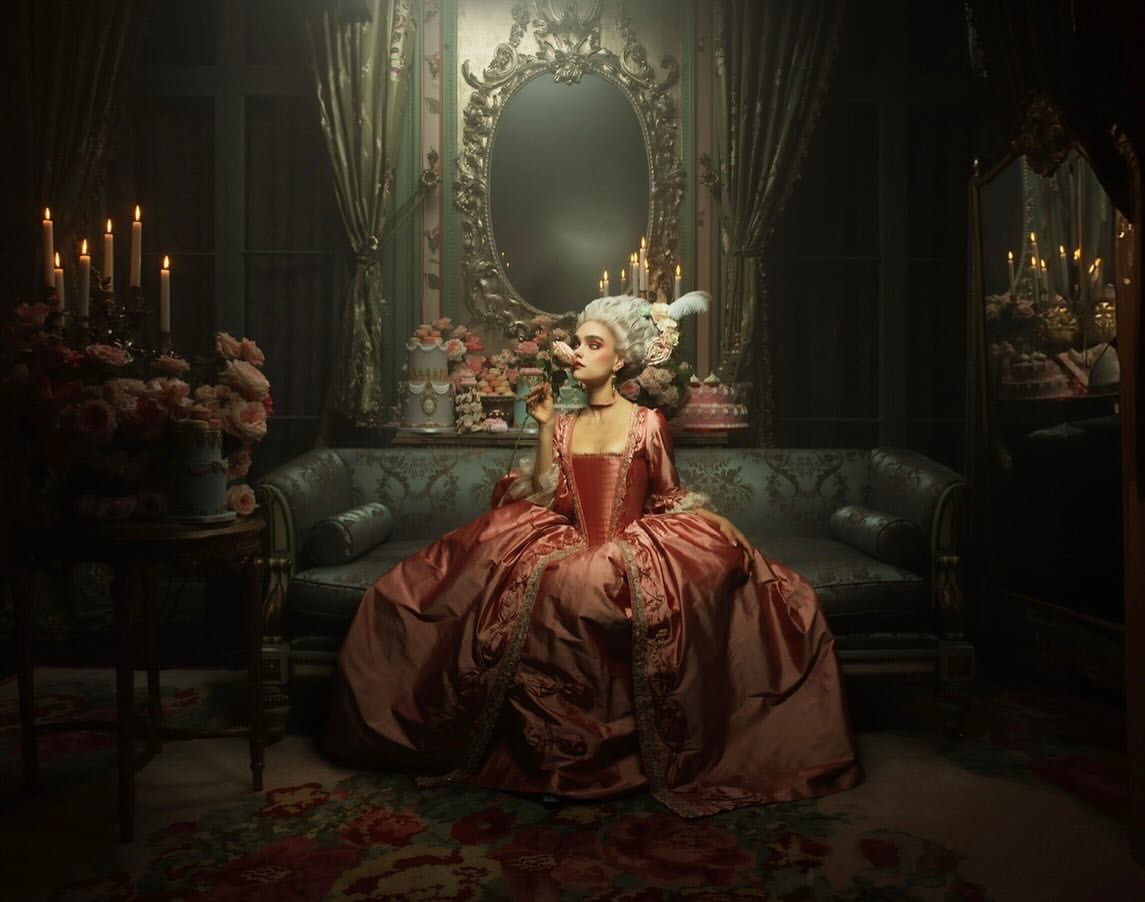 Woman in elaborate pink gown and white wig sits on a blue couch in a dimly lit room with flowers, cake and candelabras.