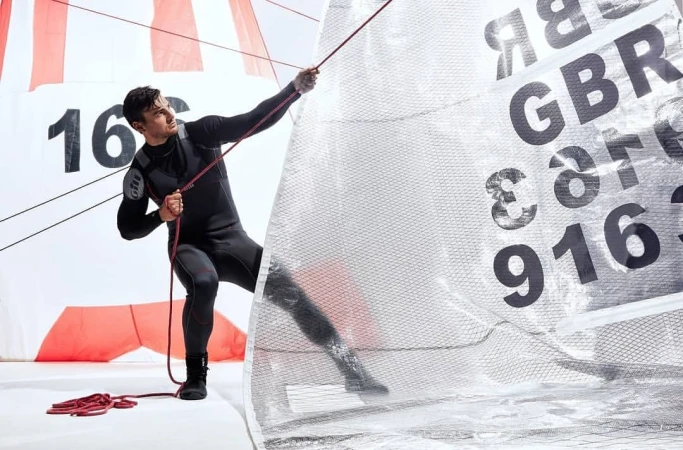 Man in wetsuit holding rope attached to sail with GBR 916 markings in a brightly lit indoor setting, showcasing sailing equipment.