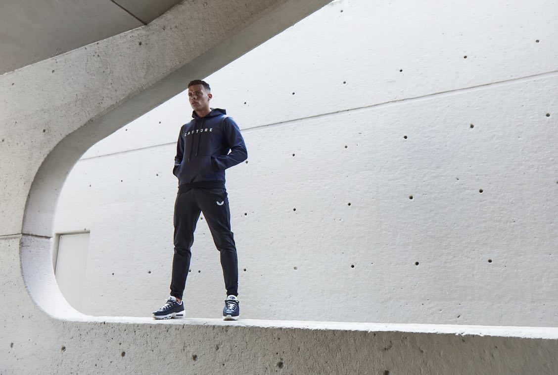 Athletic man in blue hoodie and black joggers stands confidently against a modern concrete wall with sunlight. Active sportswear fashion.