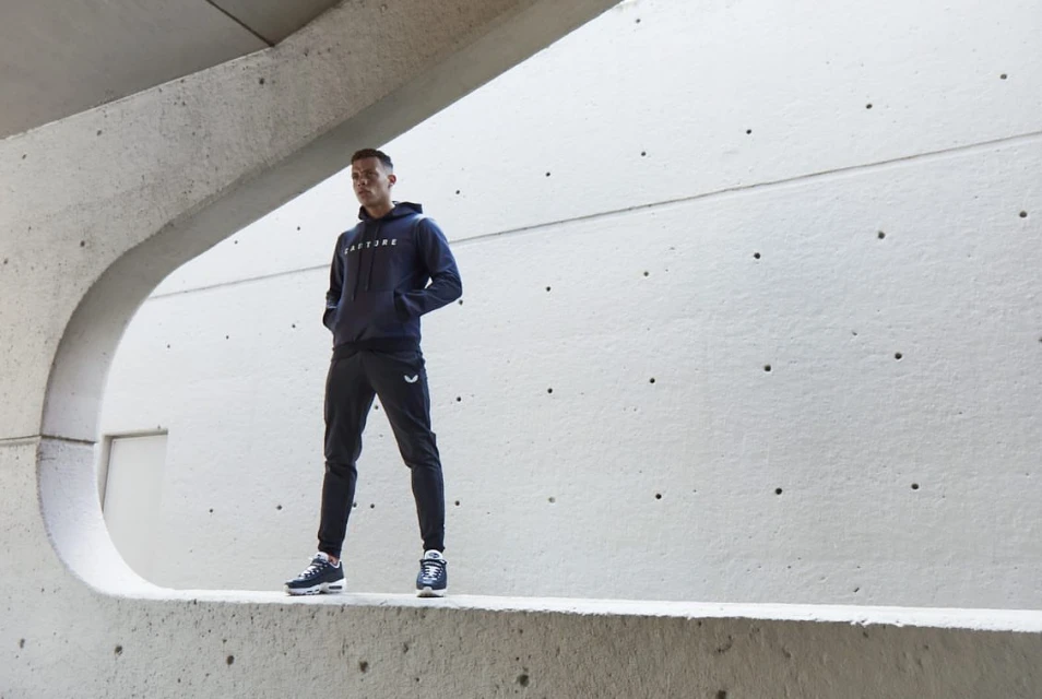 Athletic man in blue hoodie and black joggers stands confidently against a modern concrete wall with sunlight. Active sportswear fashion.