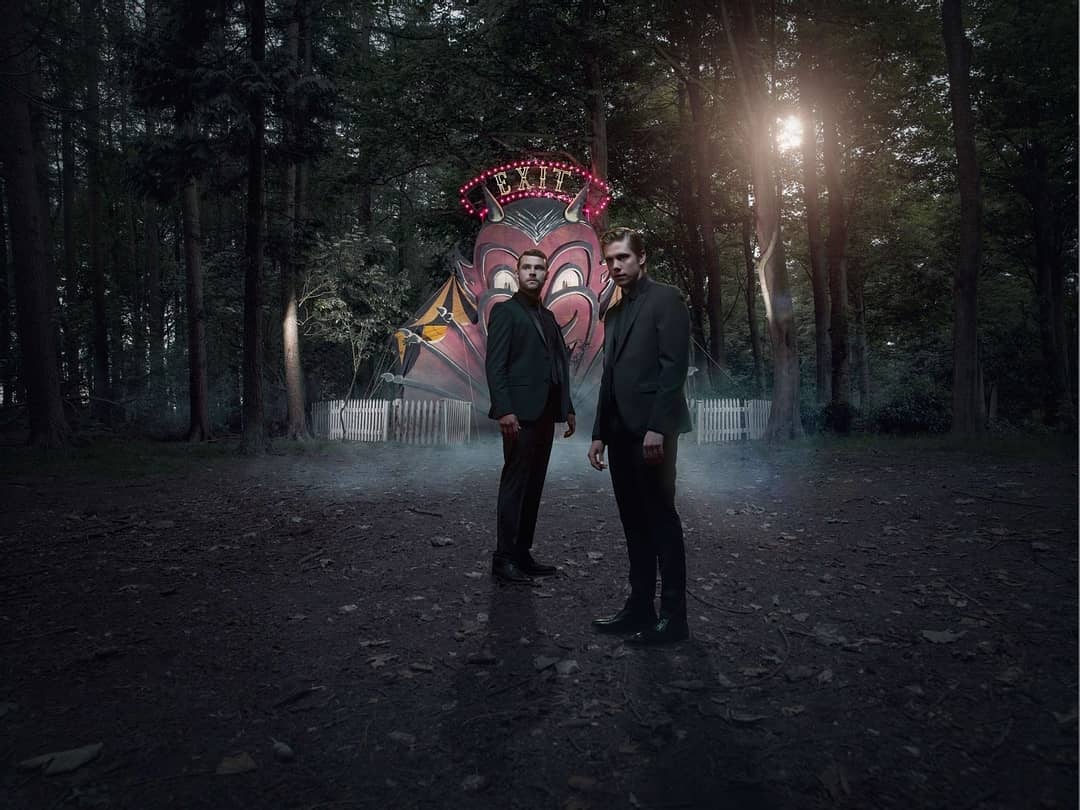 Two men in dark suits stand before a spooky carnival entrance with a devil face and 'Exit' sign in a dense, dark forest setting.