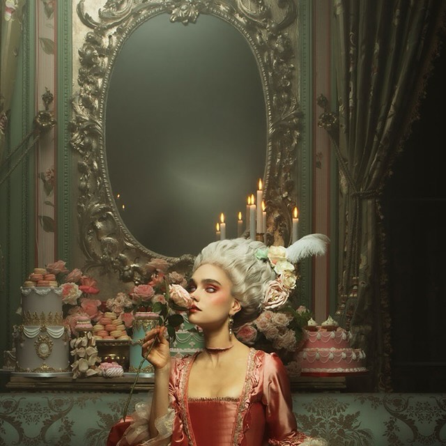 Woman in historical costume holding rose with cakes, candles, and ornate mirror in the background in an opulent room interior.