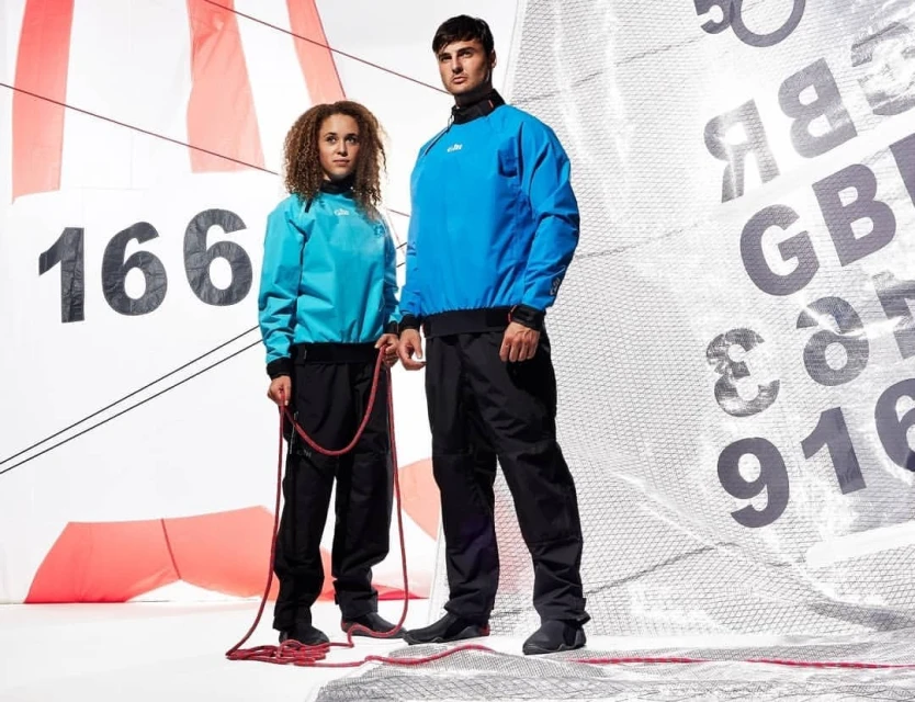 Two people in matching blue and black sailing gear stand in a photo studio with a sail backdrop holding a red rope.