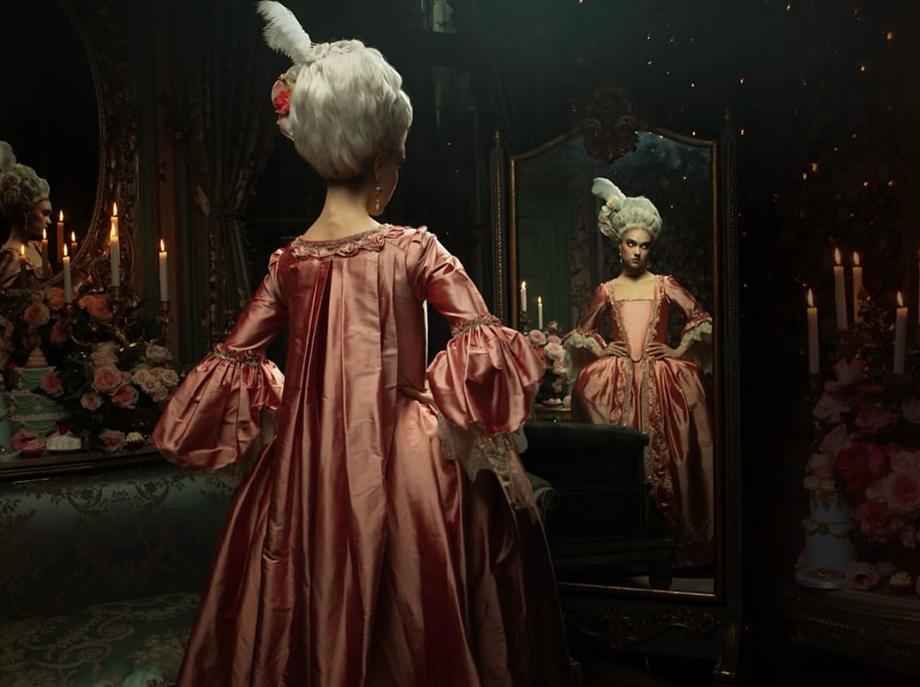 Woman in a pink, frilly dress and white wig looking in an ornate mirror in a candlelit room. The mirror reflects her image back.