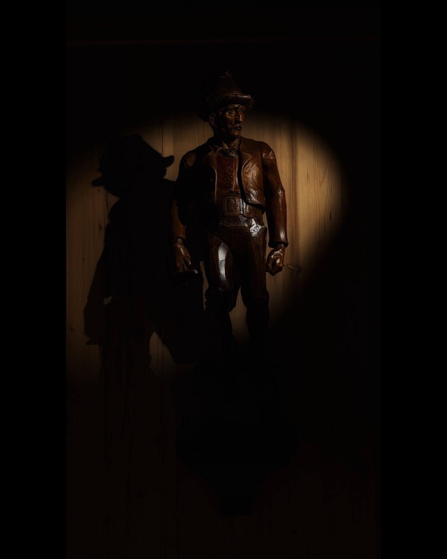 Dramatic spotlight illuminates a bronze statue of a man in a hat and jacket standing against a wooden wall casting a large shadow.