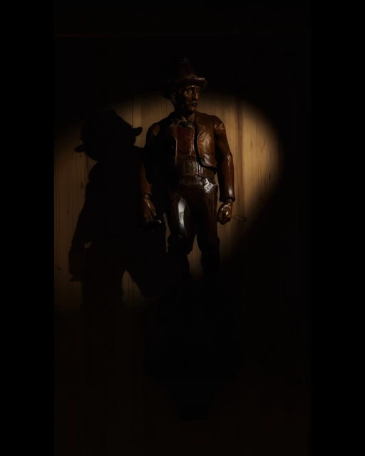 Dramatic spotlight illuminates a bronze statue of a man in a hat and jacket standing against a wooden wall casting a large shadow.