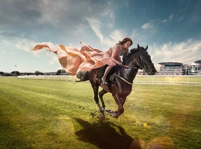 Woman in flowing gown riding a dark bay horse at full gallop on a racetrack with a large building in the background on a sunny day.