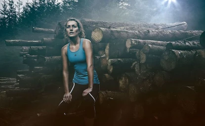 Woman in blue sports tank top and dark leggings resting against a large stack of logs in a forest.