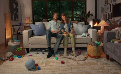 Man and woman sitting on a cream sofa holding wine glasses in a living room with toys scattered on the floor.