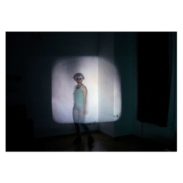 A person with short hair wearing a light green outfit is indoors in front of a wall and in the beam of light.