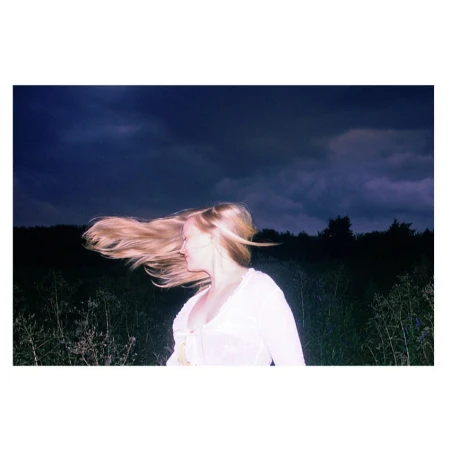 Blonde woman with hair whipping in the wind wearing a white top at night under a dark stormy sky.