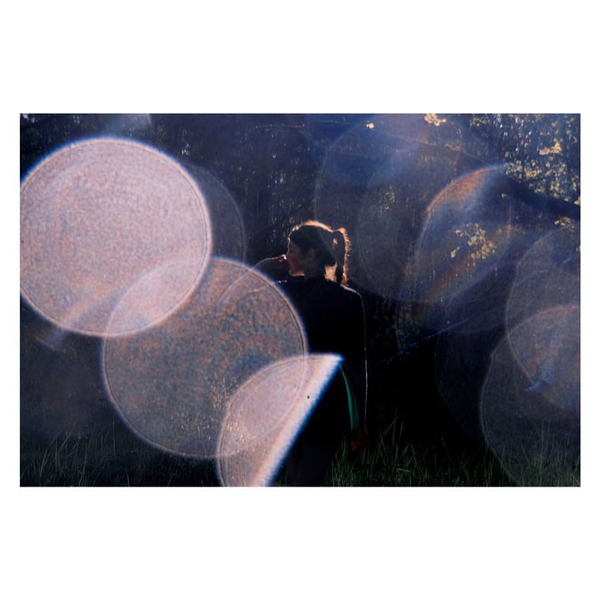Young woman stands outdoors with soft, filtered sunlight and bright bokeh orbs in foreground, set against dark foliage and natural light.