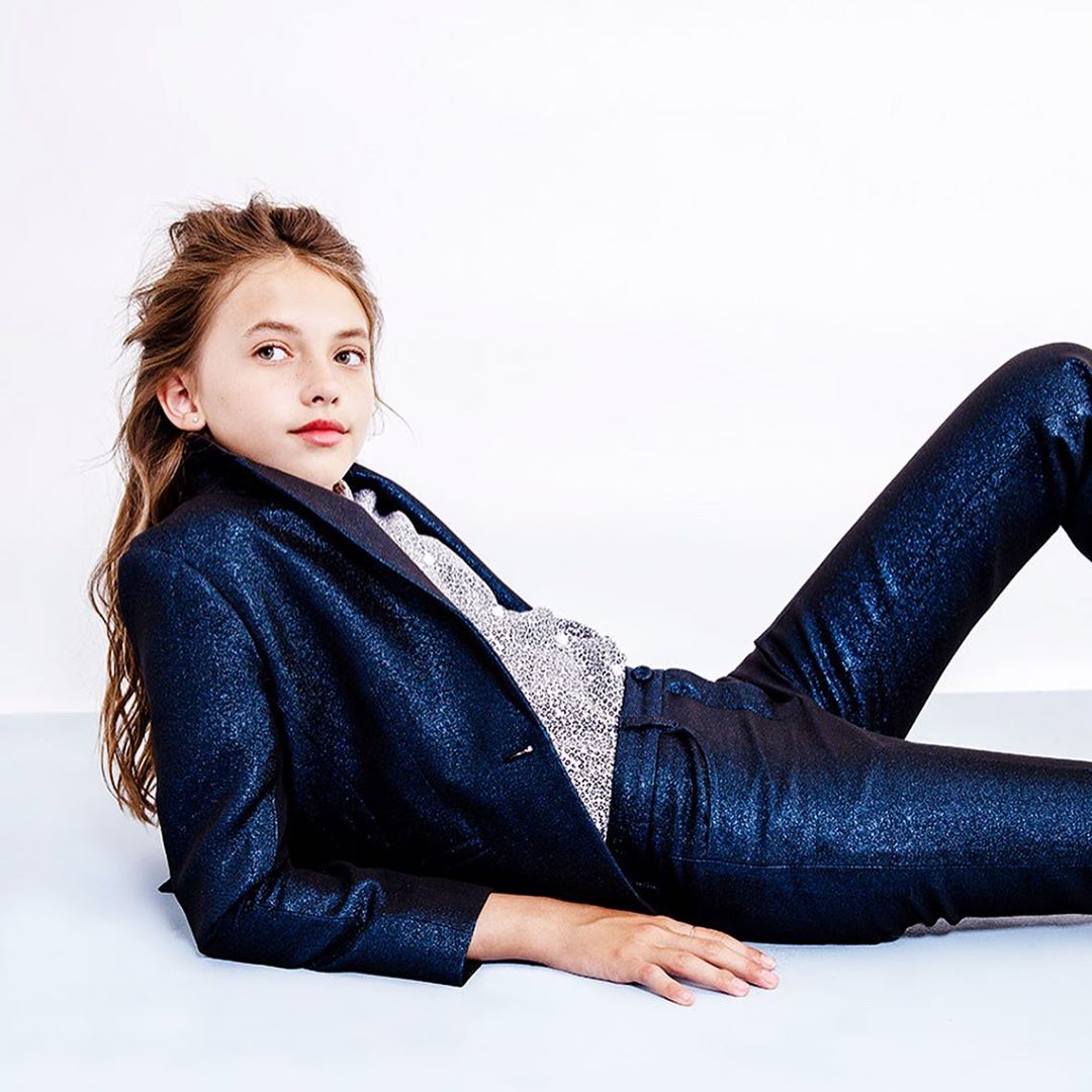 Young girl poses in a shiny dark blue suit on a white background, looking off to the side in a studio shot. Fashion photography.