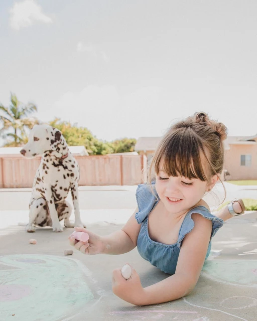 A young girl smiles while lying on concrete drawing with chalk, with a Dalmatian dog sitting in the background on a sunny day.