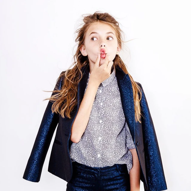 Girl in a shiny blue suit poses in front of a white wall, sticking her tongue out and placing a finger on her lips.