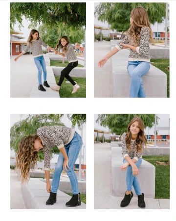 Two young girls dressed in casual sweaters and jeans posing outdoors on concrete structures, surrounded by greenery and buildings in daylight.