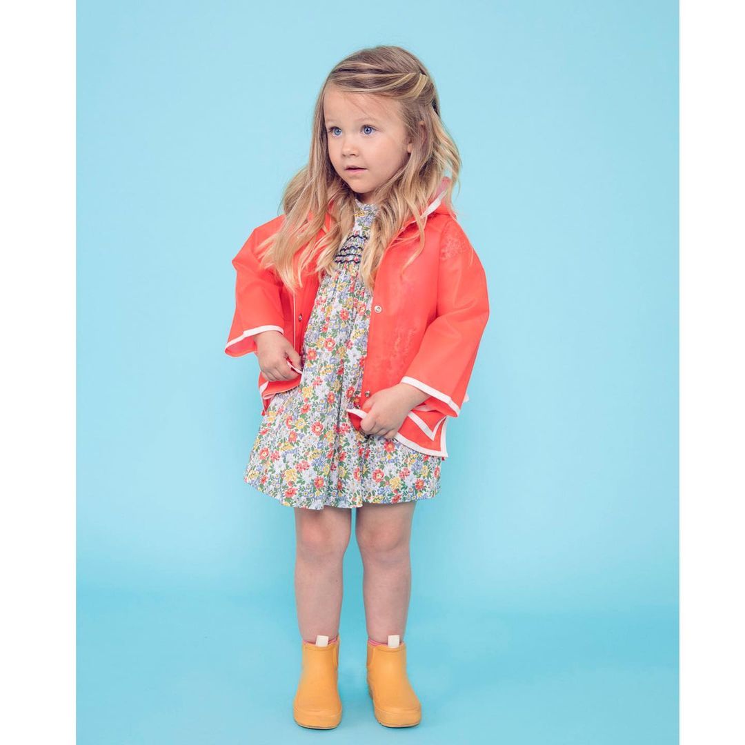 A young child stands against a bright blue background, wearing a floral dress, orange raincoat, and matching rain boots.