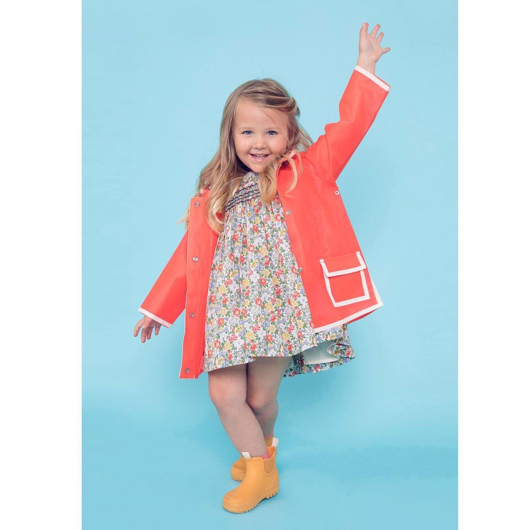 A young girl poses with a raised arm, wearing a vibrant orange raincoat, a floral dress, and yellow boots against a light blue background.